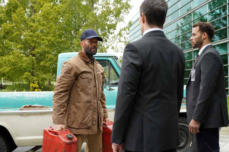 Jason Statham, com dois galões de gasolina nas mãos, olha ameaçadoramente para dois seguranças.