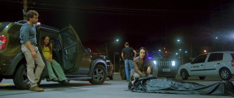 Um homem e uma mulher grávida observa a investigadora analisando um cadáver coberto na rua.