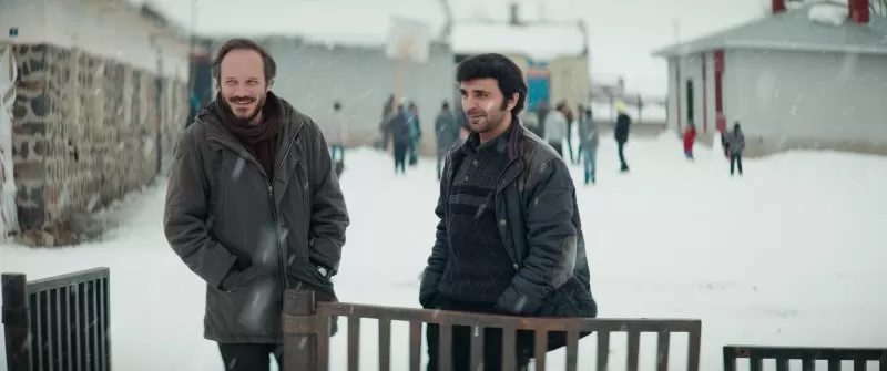 Dois homens sorridentes atrás de uma cerca na neve.