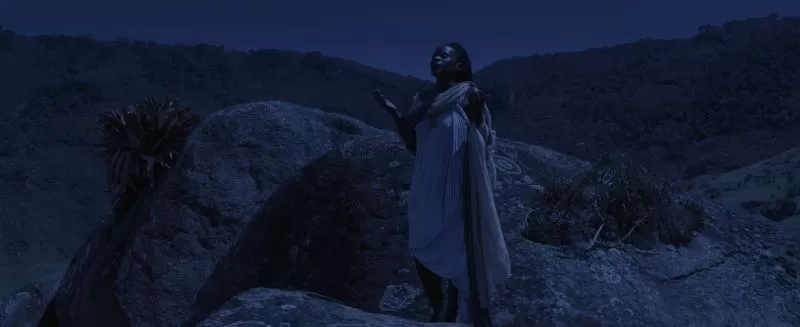 Mulher negra com vestido roxo claro de pé nas rochas à noite