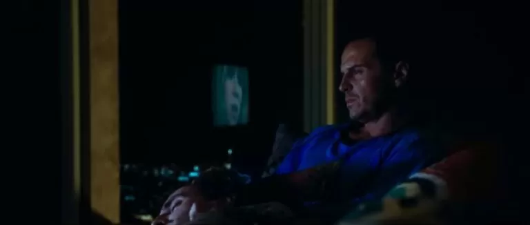 Homem sentado numa sala escura com o rosto de outro homem no seu colo.